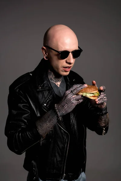 Лысый татуированный мужчина в кожаной куртке и солнцезащитных очках, держащий вкусный бургер, изолированный на сером — стоковое фото
