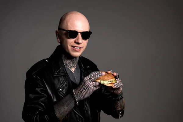 Handsome tattooed man in leather jacket and sunglasses holding hamburger and smiling at camera isolated on grey — Stock Photo