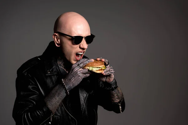 Hombre calvo tatuado en chaqueta de cuero y gafas de sol comiendo hamburguesa aislada en gris - foto de stock