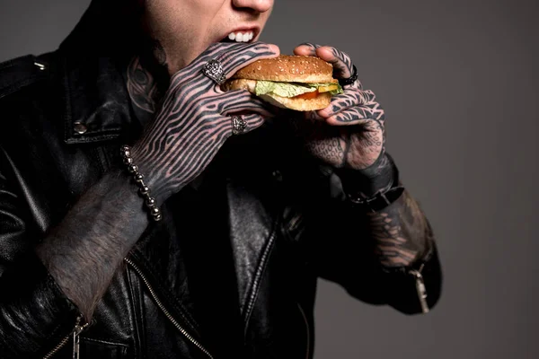 Schnappschuss eines stylischen tätowierten Mannes in Lederjacke, der Burger auf Grau isst — Stockfoto