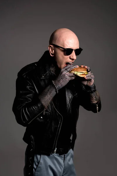 Стильный лысый татуированный мужчина в кожаной куртке и солнцезащитных очках, поедающий гамбургер изолированный на сером — стоковое фото