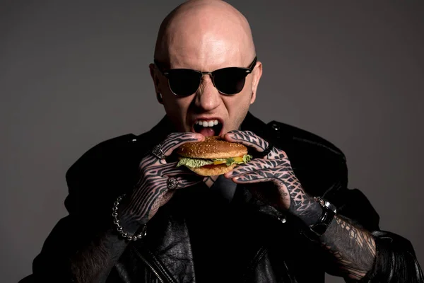 Голодний татуйований чоловік у шкіряній куртці та сонцезахисних окулярах їсть гамбургер ізольований на сірому — стокове фото