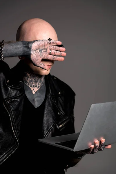 Татуйований чоловік у шкіряній куртці тримає ноутбук та ховає обличчя ізольовано на сірому — стокове фото