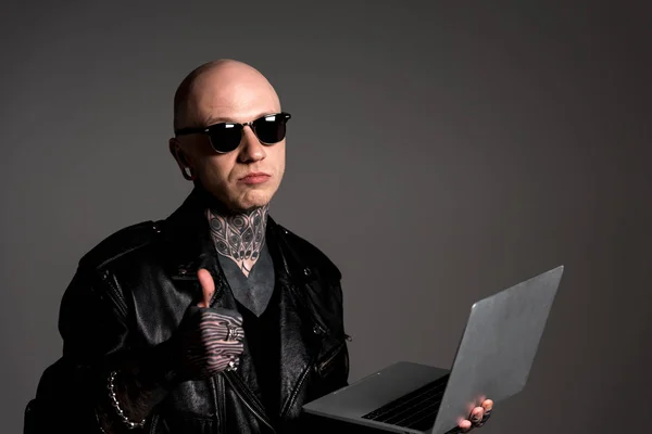 Serious tattooed man in leather jacket and sunglasses holding laptop and showing thumb up isolated on grey — Stock Photo