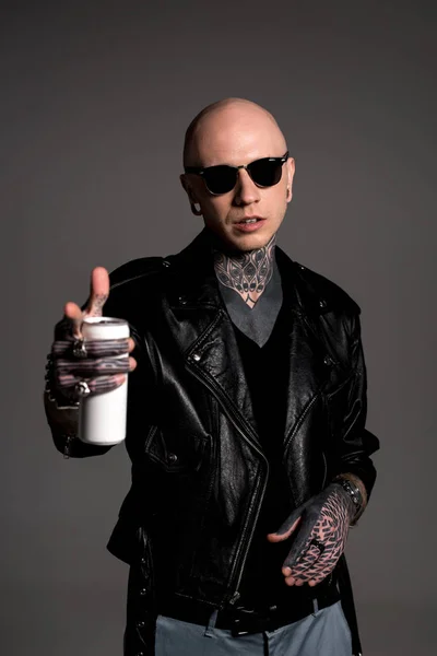 Лысый татуированный мужчина в кожаной куртке и солнцезащитных очках, держащий банку содовой и указывающий на камеру, изолированную на сером — стоковое фото