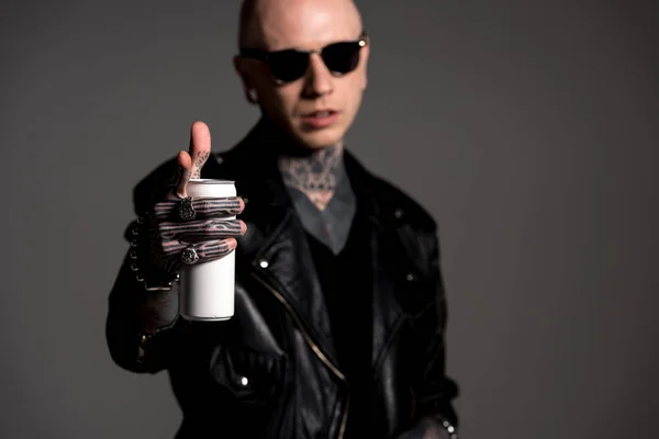 Homem tatuado elegante em jaqueta de couro e óculos de sol segurando lata de refrigerante e apontando para a câmera isolada em cinza — Fotografia de Stock