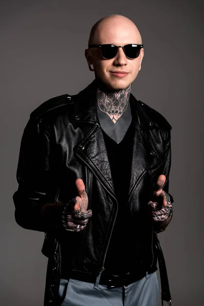 Улыбающийся татуированный мужчина в кожаной куртке и солнцезащитных очках, указывающих на камеру, изолированную на сером — стоковое фото