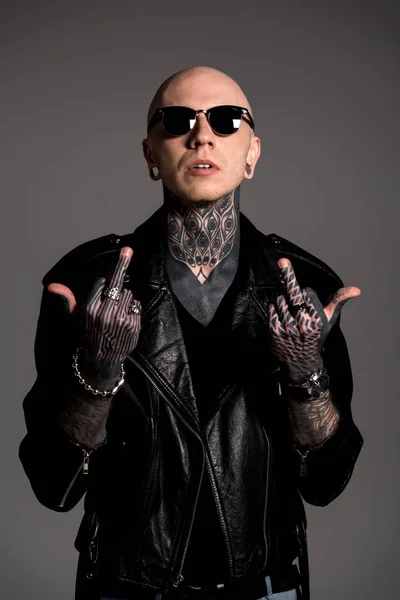 Hombre calvo tatuado en chaqueta de cuero y gafas de sol que muestran los dedos medios aislados en gris - foto de stock