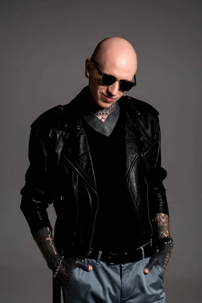 Smiling bald tattooed man in leather jacket and sunglasses standing with hands in pockets isolated on grey — Stock Photo