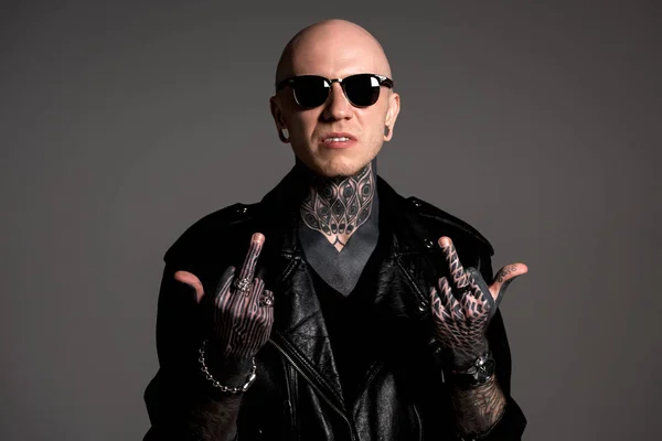 Uomo tatuato aggressivo in giacca di pelle e occhiali da sole con dita medie isolate sul grigio — Foto stock