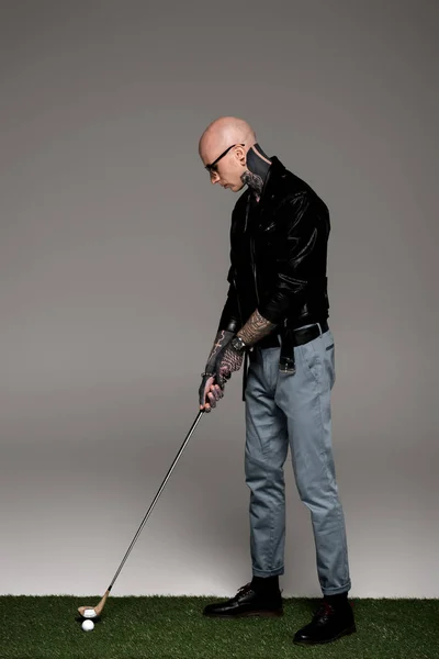 Hombre tatuado en jeans, chaqueta de cuero y gafas de sol jugando al golf en gris - foto de stock