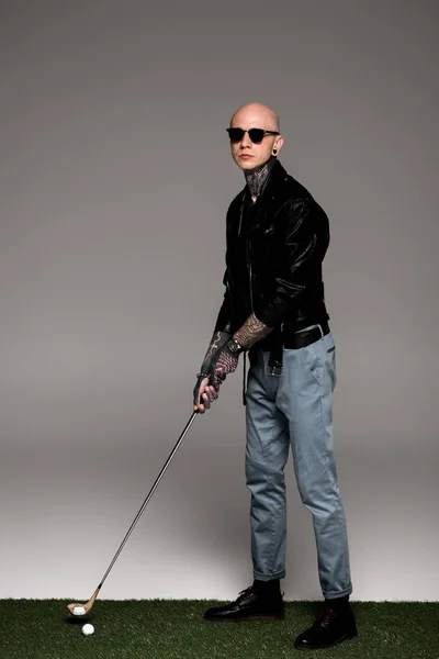 Hombre tatuado con estilo en gafas de sol y chaqueta de cuero jugando al golf en gris - foto de stock
