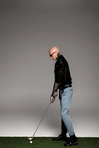 Side view of bald tattooed man in leather jacket and sunglasses playing golf on grey — Stock Photo