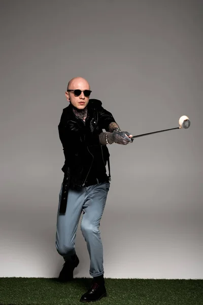 Bald tattooed man in leather jacket and sunglasses playing golf on grey — Stock Photo