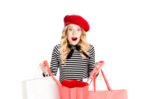 Mujer sorprendida en boina roja mirando a la cámara mientras sostiene bolsas de compras aisladas en blanco - foto de stock