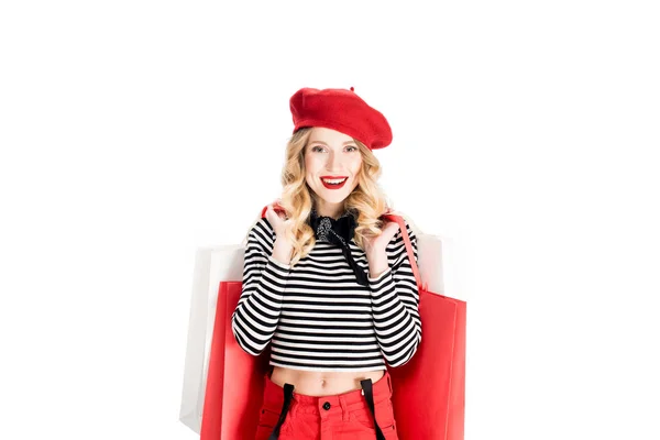 Donna allegra in berretto rosso sorridente mentre in piedi con borse della spesa isolate su bianco — Foto stock