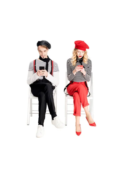 French couple in berets using smartphones while sitting on chairs isolated on white — Stock Photo