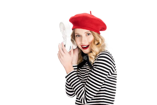 Mujer alegre en boina roja sonriendo mientras sostiene los auriculares cerca de la oreja aislada en blanco - foto de stock