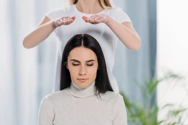 Colpo ritagliato di giovane donna con gli occhi chiusi seduto e ricevendo terapia di guarigione reiki sopra la testa — Foto stock