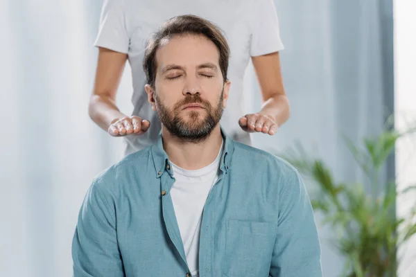 Cropped shot of bearded man with closed eyes sitting and receiving reiki healing therapy — Stock Photo