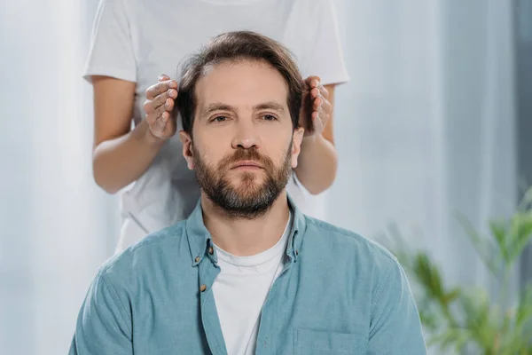 Tiro recortado del hombre barbudo sentado y recibiendo tratamiento reiki en la cabeza - foto de stock