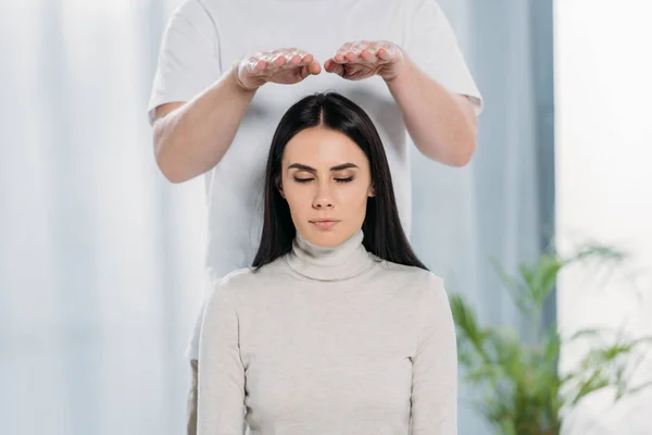 Calm young woman with closed eyes receiving reiki healing treatment above head — Stock Photo