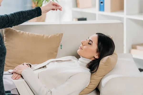 Plan recadré d'hypnotiseur avec pendule hypnotisant jeune femme couchée sur le canapé — Photo de stock