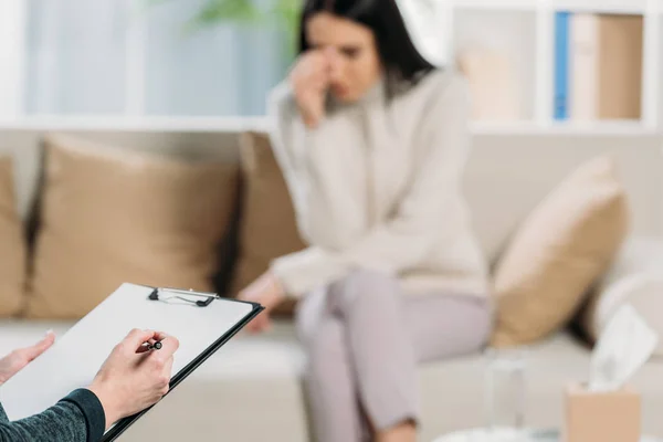 Cropped shot of psychotherapist writing on clipboard and depressed woman sitting on couch behind — Stock Photo