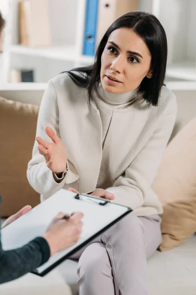 Cropped shot of psychotherapist writing in clipboard and emotional patient sitting on couch — Stock Photo