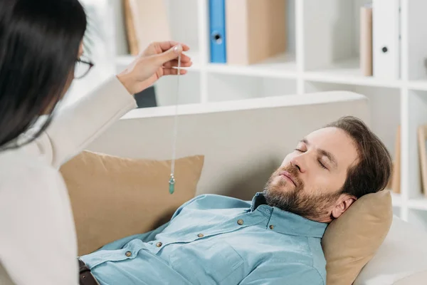 Young woman with pendulum hypnotising bearded man with closed eyes lying on couch — Stock Photo
