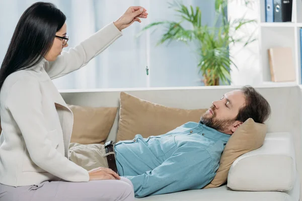 Vue latérale du jeune hypnotiseur avec pendule et homme barbu les yeux fermés couché sur le canapé pendant l'hypnothérapie — Photo de stock