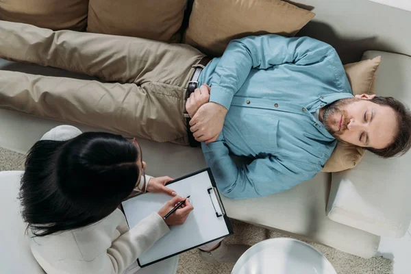 Overhead view of psychotherapist writing on clipboard and male patient with closed eyes lying on couch — Stock Photo