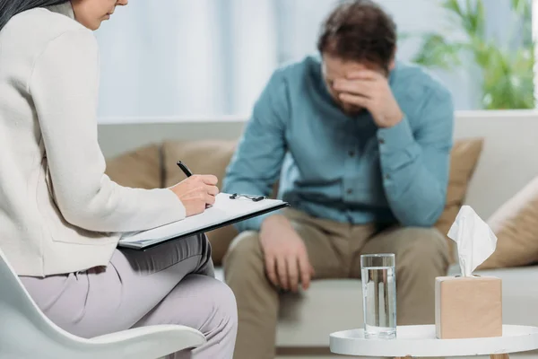 Cropped shot of psychotherapist writing on clipboard and upset patient sitting on couch — Stock Photo