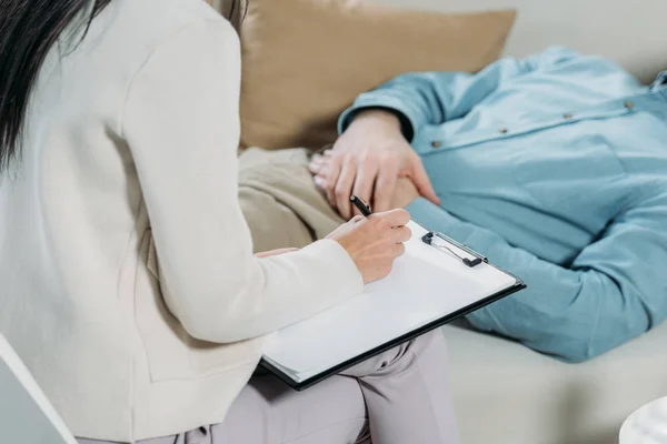Cropped shot of psychotherapist writing on clipboard and male patient lying on couch — Stock Photo
