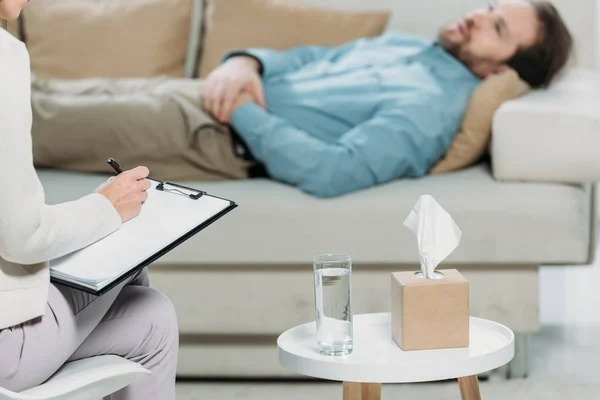 Schnappschuss eines Psychotherapeuten, der auf Klemmbrett schreibt, während ein bärtiger Mann auf der Couch liegt — Stockfoto