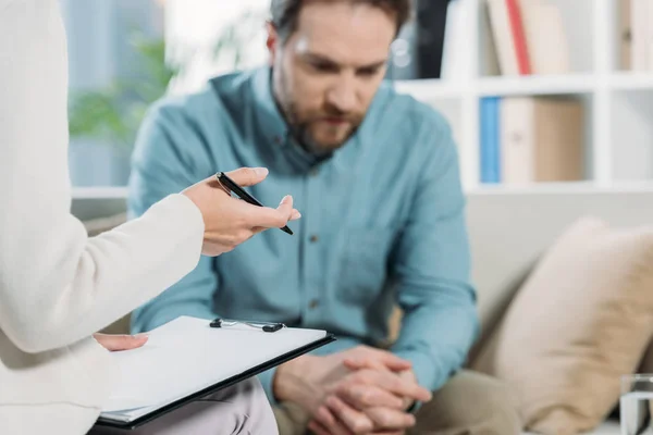 Cropped shot of psychotherapist holding pen and clipboard while upset bearded man sitting on couch — Stock Photo