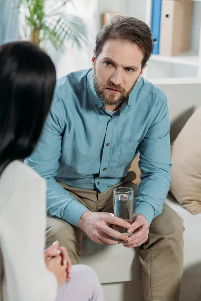 Обрезанный снимок расстроенного бородатого мужчины, держащего стакан воды и смотрящего на психотерапевта в офисе — стоковое фото