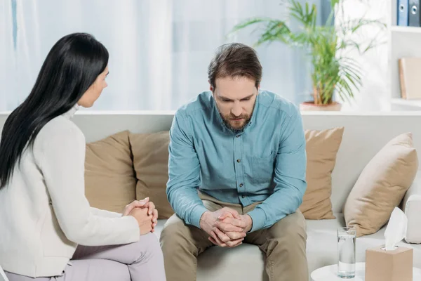 Молодой психотерапевт разговаривает с расстроенным бородатым мужчиной, сидящим на диване — стоковое фото