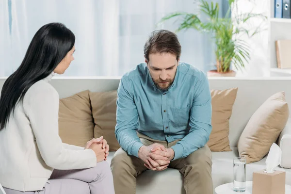 Jeune psychothérapeute parler avec bouleversé barbu homme assis sur canapé — Photo de stock