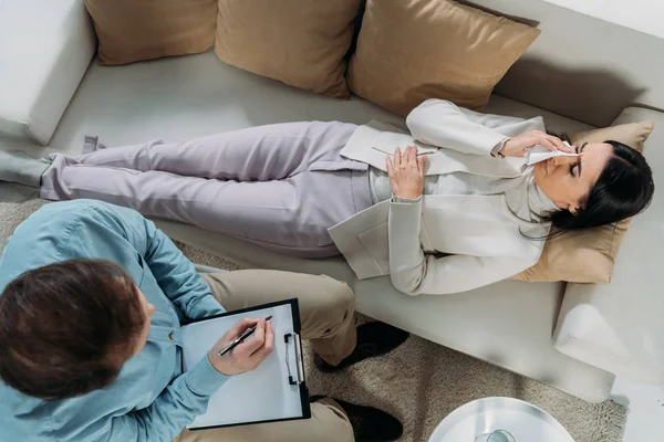 Blick von oben auf Psychotherapeutin, die auf Klemmbrett schreibt und junge Patientin weint auf Couch — Stockfoto