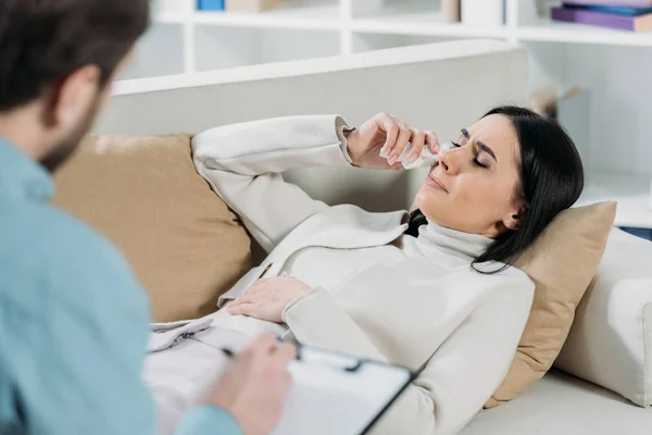 Selektiver Fokus der Psychotherapeutin auf Klemmbrett und junge Patientin weint auf Couch — Stockfoto