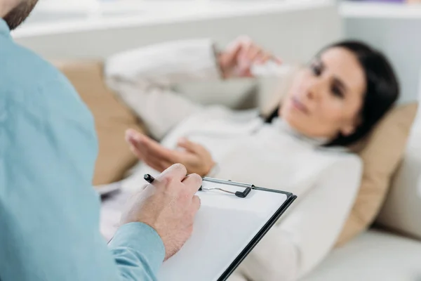 Cropped shot of psychotherapist writing on clipboard and young patient in depression lying on couch — Stock Photo