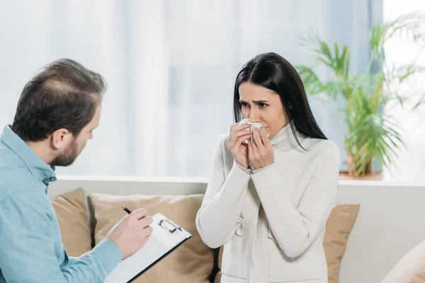 Aufgebrachte junge Frau weint und schaut bärtigen Psychotherapeuten mit Klemmbrett an — Stockfoto