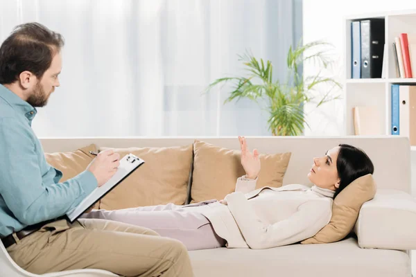 Vista laterale della barba psicoterapeuta scrittura sugli appunti e giovane paziente sdraiato sul divano — Foto stock