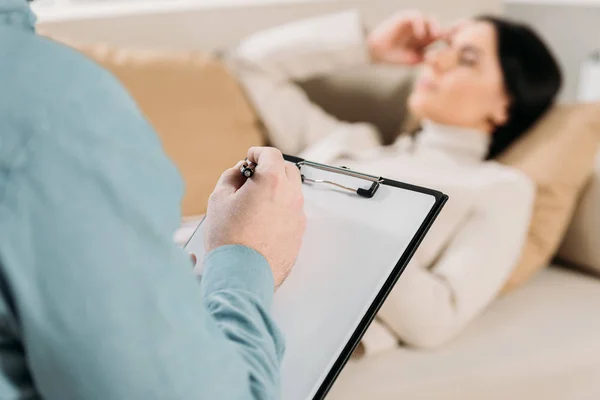 Recortado disparo de psicoterapeuta escritura en portapapeles y joven mujer acostada en el sofá en la oficina - foto de stock