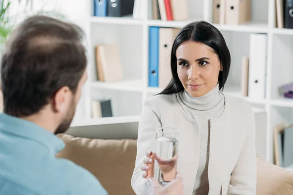 Giovane donna in possesso di un bicchiere d'acqua e guardando psicoterapeuta in ufficio — Foto stock