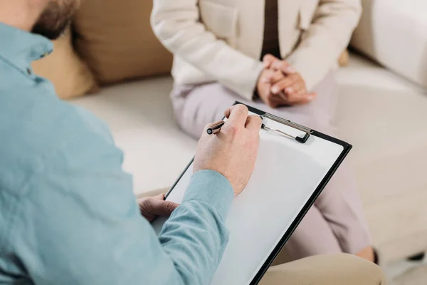 Cropped shot of psychotherapist writing on clipboard and female patient sitting on couch — Stock Photo