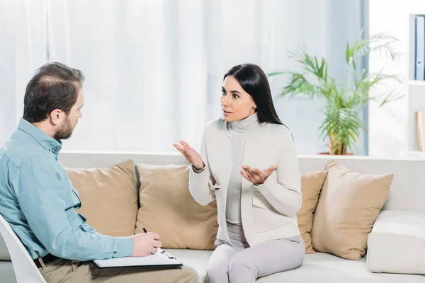 Emotionale Patientin spricht und schaut Psychotherapeutin beim Schreiben auf Klemmbrett an — Stockfoto