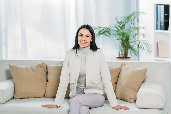 Attractive young woman sitting on sofa and smiling at camera — Stock Photo