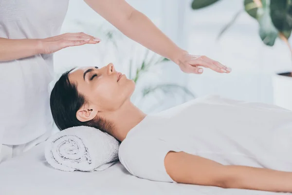 Tiro recortado de mujer joven con los ojos cerrados que reciben tratamiento de reiki en la cabeza y el pecho - foto de stock